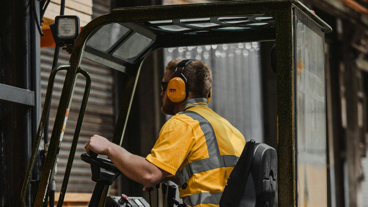 Operator in hi-vis and ear defenders driving a counterbalance lift truck in a loading bay